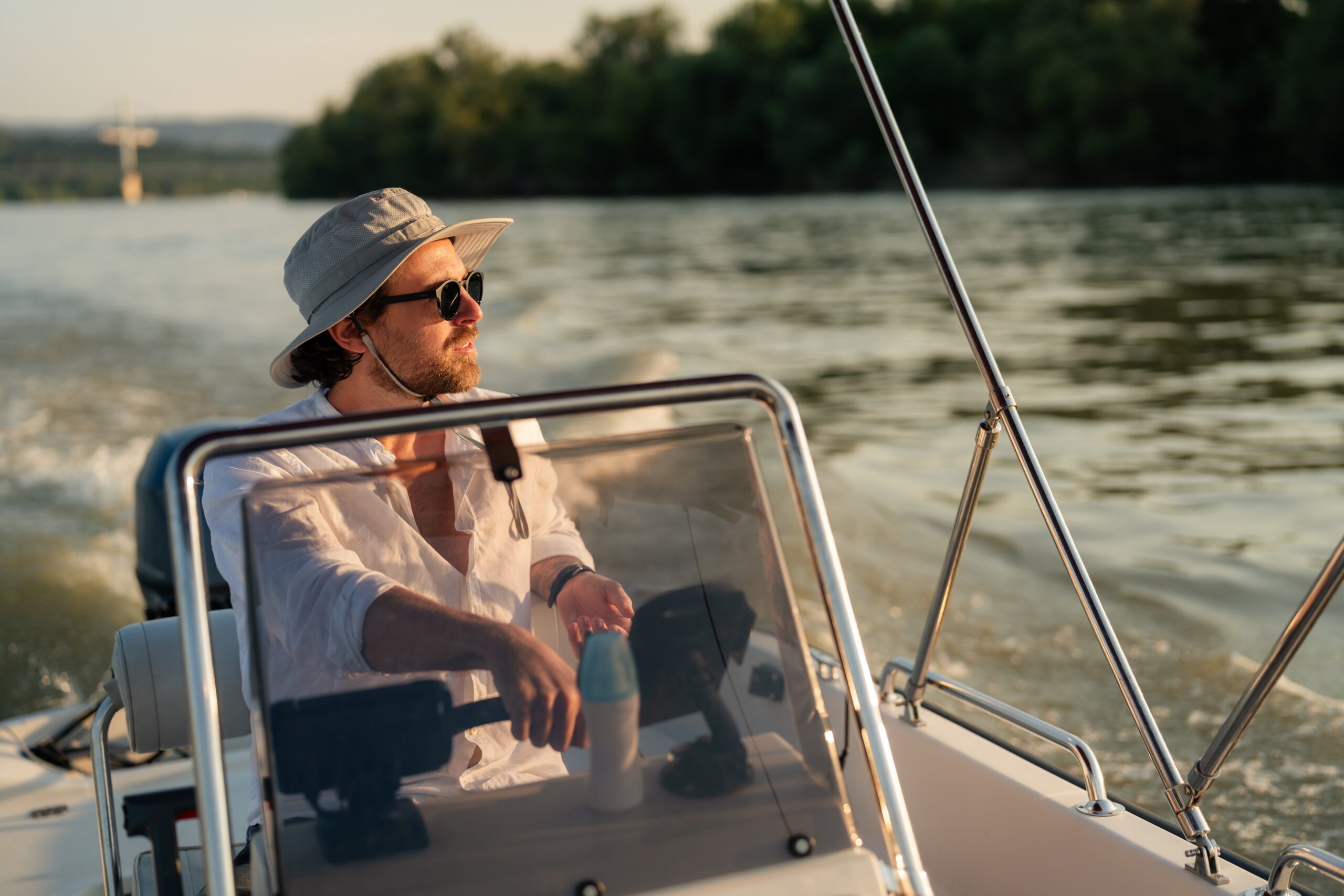 man behind the helm of a boat wearing a hat, sunglasses, and long sleeve shirt. 
